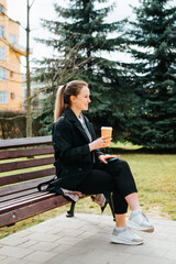 Wall Mural - Charming positive young woman in stylish coat resting on bench during coffee break outdoors. Side view of smiling pretty lady holding disposable coffee cup and looking away in park