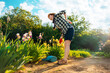 © _KUBE_ - Caucasian adult woman stands bend over near a flowering iris bush, holding her back in pain. A watering can is lying on the ground. Bottom view. Concept of back health problems