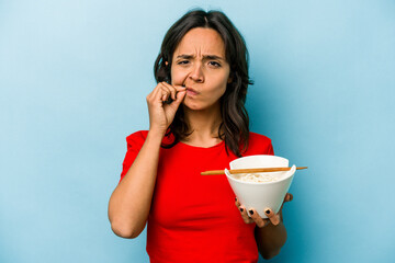 Wall Mural - Young hispanic woman eating noodles isolated on blue background with fingers on lips keeping a secret.