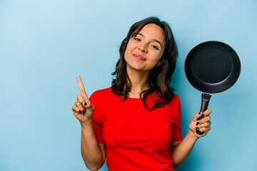 Wall Mural - Young hispanic woman holding a frying pan isolated on blue background showing number one with finger.