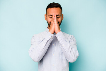 Young hispanic man isolated on blue background holding hands in pray near mouth, feels confident.