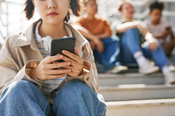 closeup of teenage girl holding smartphone outdoors while sitting on metal stairs with group of frie