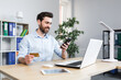 © Liubomir - Successful online shopping through a smartphone. Successful deal. Satisfied young man holds a credit card and a mobile phone, makes an order. Sitting at a desk in the office on a laptop