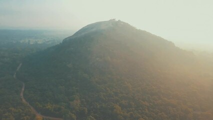 Wall Mural - Drone panning above point of interest Sigiriya Lion Rock in Sri Lanka at sunrise with bright light mist and haze filled skies overlooking natural tropical forest with orange blue tones