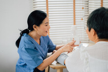 Happy Asian Female Caregiver Smiling And Pointing At Bottle Of Pills While Sitting At Table And Measuring Blood Pressure Of Senior Woman Against Window At Home