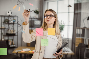 Caucasian woman in glasses holding digital tablet and drawing financial graphic on glass wall before office meeting. Female administrative manager preparing for presentation at work.