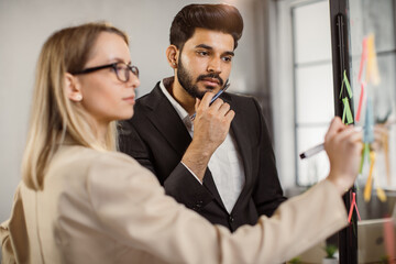 Focused indian employee in elegant suit pensively watching his caucasian colleague making notes on stickers attached to glass wall in office room. Business strategy and cooperation concept.