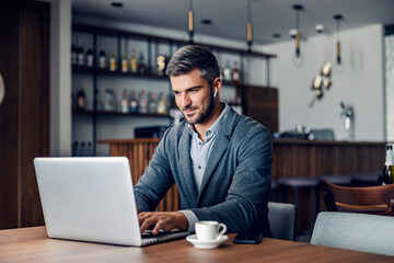 A dedicated busy man dressed in smart casual is sitting in working friendly cafe and typing a report on the laptop. He has earphones in his ears. A businessman typing on a laptop in a cafe.