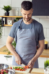 Wall Mural - Handsome father, strong young man cooking healthy vegetable salad with fresh organic ingredients, tasty food in the kitchen at home . Men doing chores. Ripe pepper, tomato, cucumber