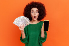 Portrait Of Shocked Woman With Afro Hairstyle Wearing Green Casual Sweater Holding Fan Of Hundred Dollars Bills And Cell Phone With Empty Display. Indoor Studio Shot Isolated On Orange Background.