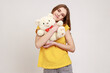 Portrait of happy pleasant looking teenager girl wearing yellow T-shirt hugging romantic present white soft Teddy bear, looks satisfied. Indoor studio shot isolated on gray background.
