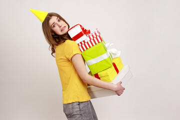 Wall Mural - Funny woman of young age with brown hair and party cone on head holding lot of gift boxes, embracing many heavy birthday presents, wearing T-shirt. Indoor studio shot isolated on gray background.