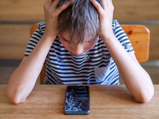 Portrait of scared kid girl with broken mobile phone on table. Sad child broke screen of smartphone. Teen boy in desperation is holding his head and looking at phone with cracked screen at day time