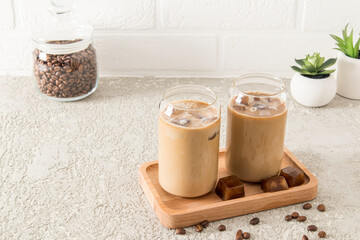 two modern glasses in the shape of a can of cold latte coffee, a chocolate drink or mocha on a wooden tray opposite a white brick wall.