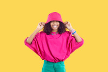 Happy Joyful Kid In Trendy Casual Outfit Puts Pink Bucket Hat On Her Head. Cheerful Beautiful Black Girl Wearing Loose Fuchsia Top And Green Pants Having Fun And Laughing. Children's Fashion Concept