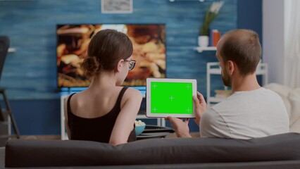 Wall Mural - Man holding digital tablet with green screen watching online video content with girlfriend sitting on sofa. Couple looking at touchscreen device with chroma key enjoying influencer vlog.