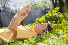 Spring Awakening. Slow Life. Enjoying The Little Things. Dreaming Of Spring. Middle Aged Woman Uses Smartphone Lying On The Grass With Dandelions On A Spring Day.. Social Networks