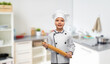 cooking, culinary and profession concept - happy smiling little girl in chef's toque and jacket with rolling pin over home kitchen background