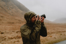 Male Photographer In Hooded Jacket Using Camera Below Mountain, Glen Etive, Scottish Highlands, Scot
