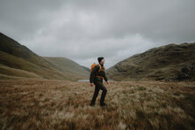 Male Hiker With Backpack Hiking Below Hills In Remote Landscape, Great End, England
