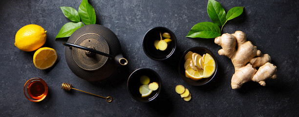 Wall Mural - Ginger, lemon, honey tea in black cups , tea pot with fresh slices, roots. Dark background. Top view.