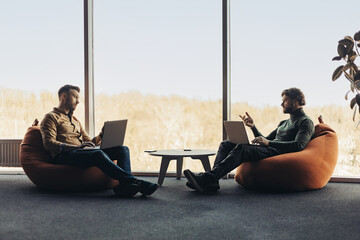 Poster - Young colleagues sitting in bean bag chairs, working with laptops, communicating with each other at open space office
