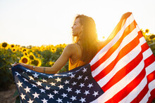 Young Woman Proudly Hold Waving American USA Flag In In The Sunflower Field. Patriot Raise National American Flag At Sunset. Independence Day, 4th July.
