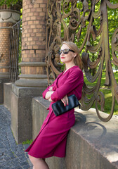 A beautiful blonde 30 years old in a red dress stands near a beautiful fence, in a park, during the day. City tour, sales, tourist season, stylish image of a girl