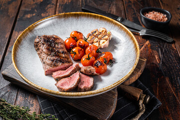Roasted Lamb tenderloin meat in plate with grilled tomato and garlic, mutton sirloin fillet steak. Dark background. Top view