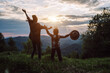 Back view of young mother and little daughter kid watching sunset in evening at camping. Caring mom with small girl child jumping on mountains background. Family activity adventure on vacation concept