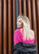 Happy beautiful young girl with a cute smile and blond hair in a fashionable knitted pink sweater walks in the city near the metal wall. Natural Female Positive Emotions