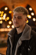 Fresh male indoors portrait of young handsome man with hairy in trendy clothes with jacket strands and looks at the camera in mall with lights bokeh