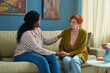 © pressmaster - African american female social worker talking to elderly woman during her visit while they sitting on sofa in living room