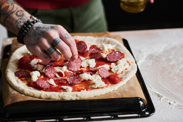 Close-up shot of unrecognizable man with tattooed arms cooking pizza at home adding salami slices