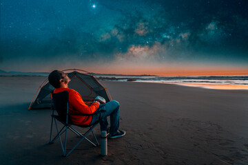 Wall Mural - man sitting in a camping chair alone on the beach looking at the starry night next to a tent