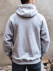 Back view of young man in a hoodie with hood for advertising with ability to apply logo. A man stands on a background of light gray concrete wall. Design and layout of men's uniforms for printing.