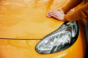 The girl folded her hands on the hood of her yellow sports car. Modern optics on the background of a beautiful car. A young woman near a close-up of the car.