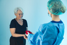 Female Doctor In Medical Uniform, Mask And Medical Cap Giving An Artificial Human Heart To An Old Lady With Grey Hair Wearing A Black Outfit And Glasses. Studio Shot. High Quality Photo