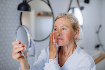 Poster - Beautiful senior woman in bathrobe looking at mirror and applying natural face cream in bathroom, skin care concept.