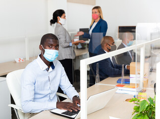 Wall Mural - Portrait of focused afro american businessman in medical face mask working in open plan office. Pandemic prevention and social distancing concept