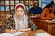 © pressmaster - Young adult immigrant Muslim woman wearing hijab sitting at desk in classroom making notes during lesson