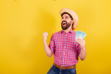 Bearded man wearing typical clothes for party Junina. Running and celebrating holding money banknotes, victory. For the Arraia Party