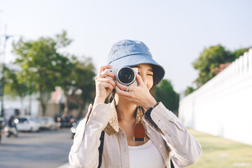 Wall Mural - Attractive happy smile young adult asian woman traveller wear blue hat and backpack. Using camera for photo trip. People traveling in city lifestyle on day. Staycation summer trip concept.