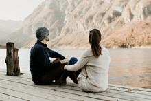 A Couple In Love Is Sitting On A Wooden Pier By A Lake In Europe, In The Alps. They Sit Next To Each Other, Facing The Distance. Romantic Sunset Date Scene. Travel Destination Idea, Relaxing Scenery.