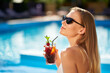 © artiemedvedev - Blonde woman drinks refreshing cocktail sunbathing and sitting near swimming pool at tropical spa. Female in sunglasses enjoys a drink on poolside. Girl chilling in tropical resort on vacation.