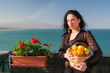 View of a typical mediterranean woman holding a wicker basket full of oranges and lemons in a panoramic balcony in Sicily, with blue sea and Mount Etna in the background