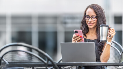 Wall Mural - Good-looking woman drinks coffee from reusable mug and checks text messages on a phone with an open laptop in front of her