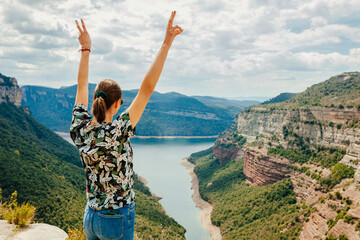 Wall Mural - Rear view of young hiker female standing with arms outstretched in peace sign gesture, enjoying on top rocky mountains. Adventure, landscape, holiday concept.