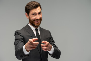 Positive optimistic bearded man pointing fingers at camera and looking with toothy smile, believes in you, motivation, wearing official style suit. Indoor studio shot isolated on grey background