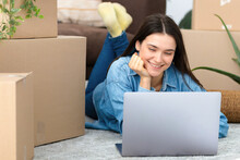 Moving To A New House, Rental Housing. Happy Young Caucasian Woman Using Laptop Computer To Search And Order A Transportation Service And Movers To Move To A New Home